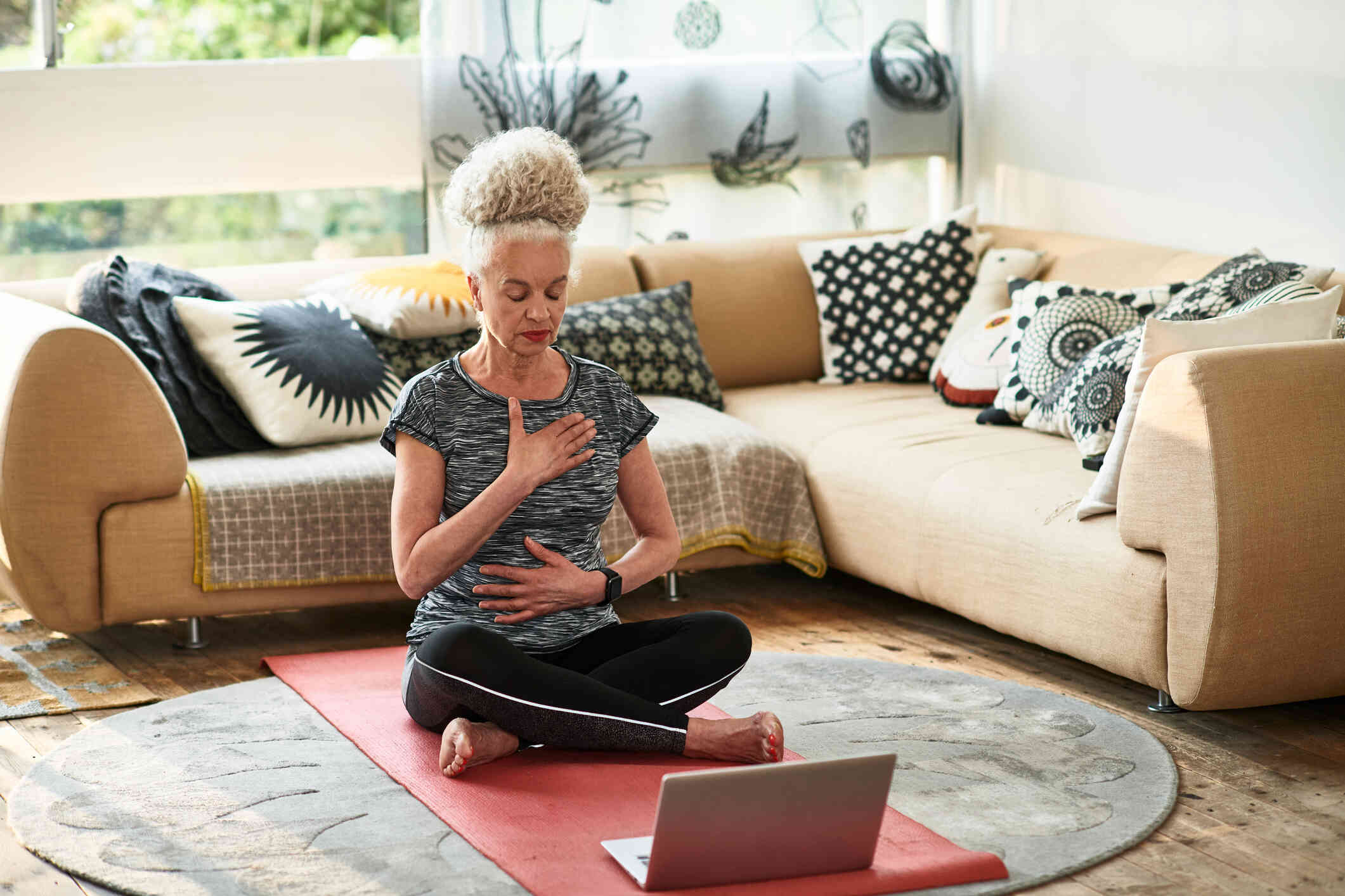 A woman sits on a yoga mat on the floor of her living room with her eyes closed as she meditiates.